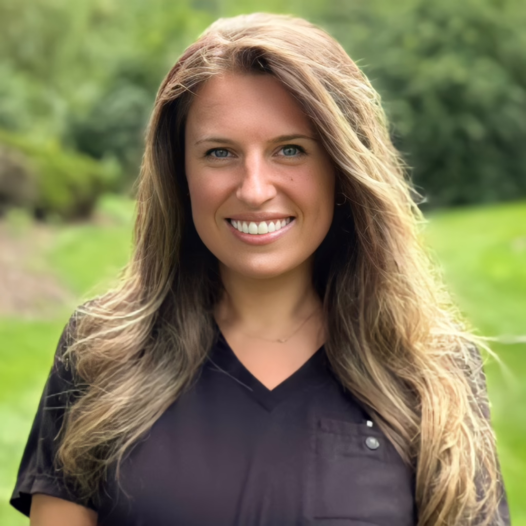 Portrait of a smiling woman with long light brown hair outdoors against a green, blurred background.